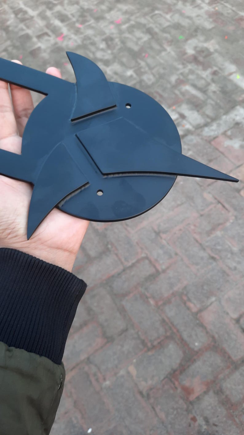 May include: A black, geometric metal sculpture with a circular base and angular, pointed extensions. The design features sharp lines and a symmetrical arrangement, held in a hand against a brick background.