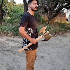 Puede incluir: Un hacha de batalla de doble filo con una cabeza de metal color bronce con dos hojas curvas y un mango de madera largo y recto. El hacha se sostiene horizontalmente, mostrando el contraste entre las hojas de metal pulido y la veta de madera clara del mango, que se extiende desde el punto de montaje central.