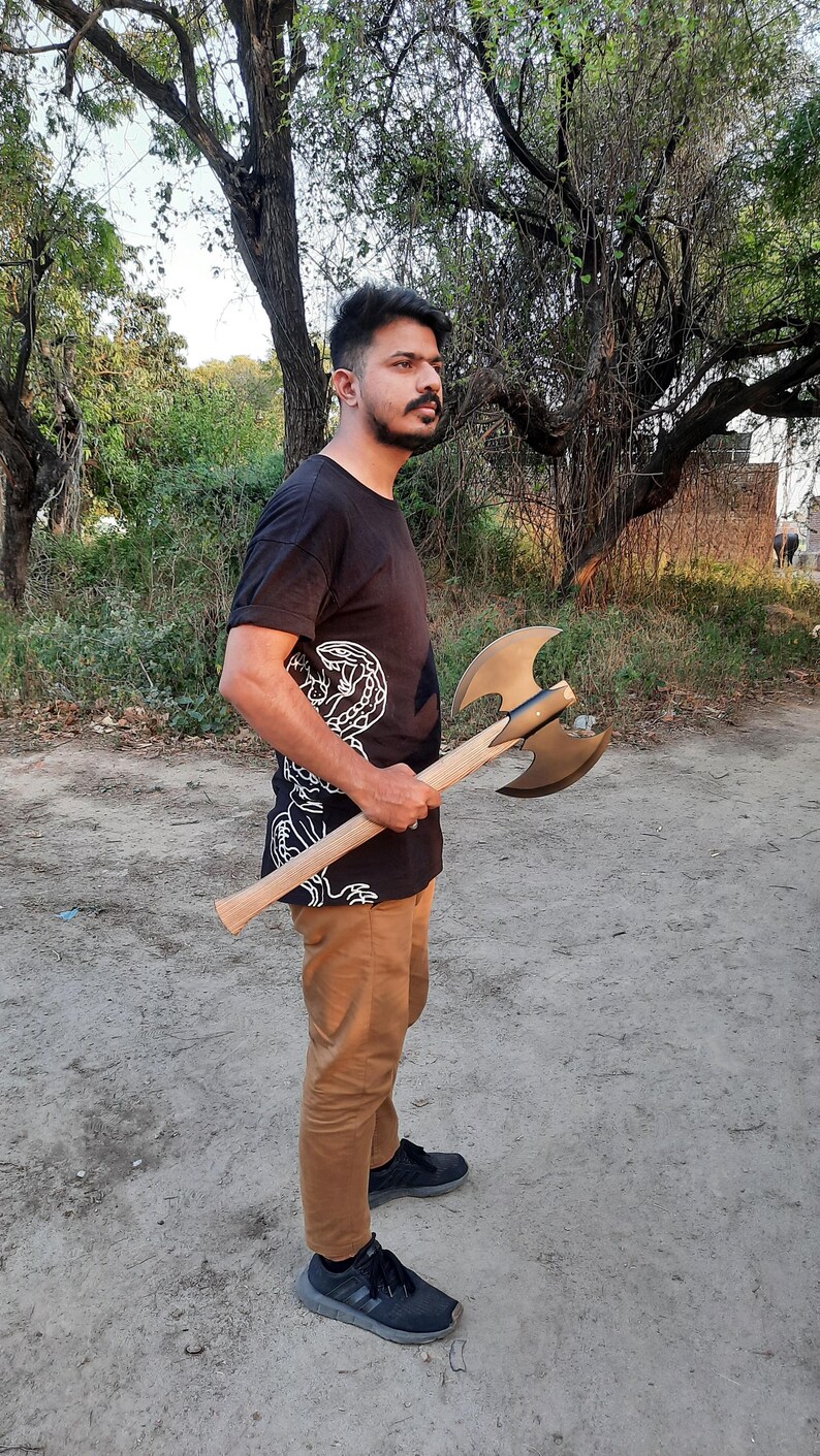 Puede incluir: Un hacha de batalla de doble filo con una cabeza de metal color bronce con dos hojas curvas y un mango de madera largo y recto. El hacha se sostiene horizontalmente, mostrando el contraste entre las hojas de metal pulido y la veta de madera clara del mango, que se extiende desde el punto de montaje central.