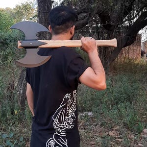 Puede incluir: Un hacha de batalla de doble filo con una cabeza de metal negro mate y un mango de madera clara. El hacha se sostiene horizontalmente sobre los hombros. El mango presenta una textura de veta de madera visible y un extremo ensanchado.