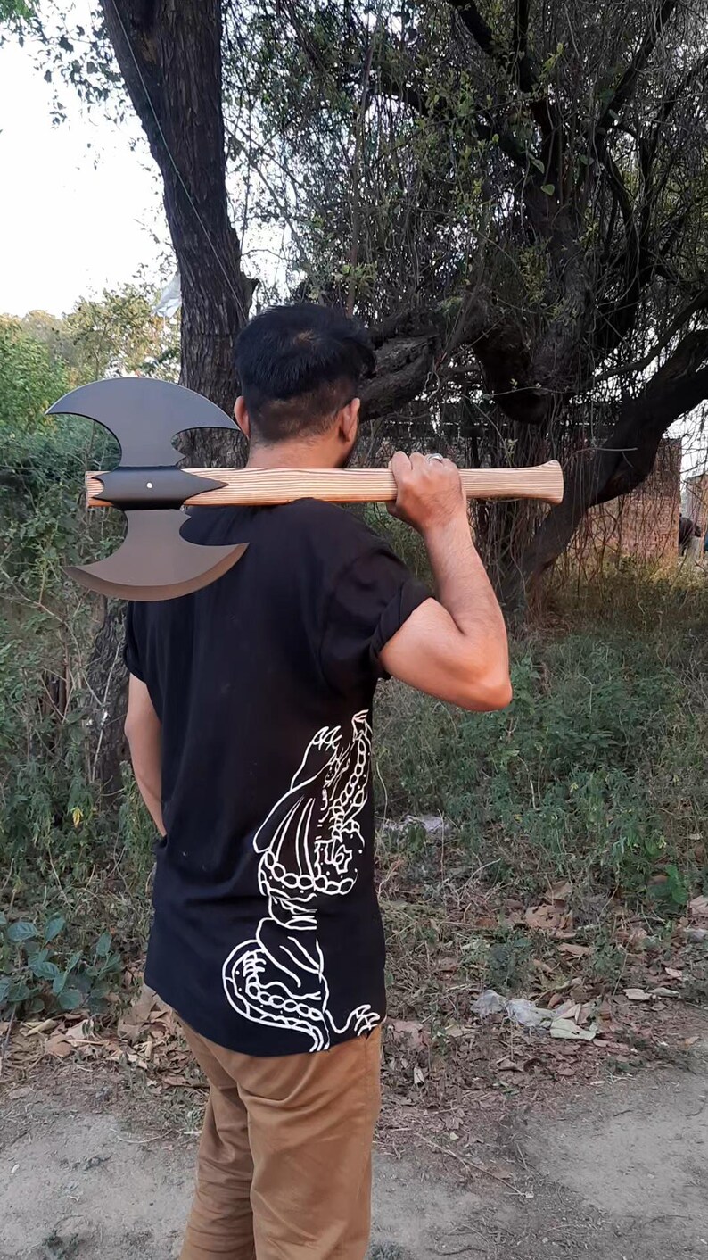 Puede incluir: Un hacha de batalla de doble filo con una cabeza de metal negro mate y un mango de madera clara. El hacha se sostiene horizontalmente sobre los hombros. El mango presenta una textura de veta de madera visible y un extremo ensanchado.