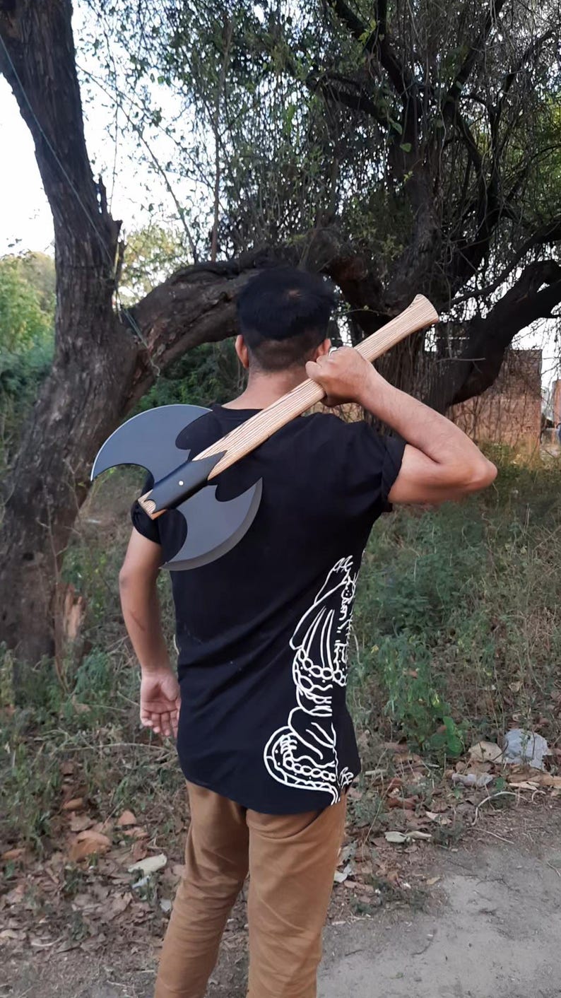 Puede incluir: Un hacha de batalla de doble filo con una cabeza de metal negro mate con dos hojas curvas y un mango largo de madera clara. El hacha se sostiene sobre el hombro, mostrando el contraste entre el metal oscuro y la veta de la madera natural.