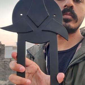 May include: A black metal decorative object with a geometric design. The object has a circular top with a star-like shape and two long, parallel bars extending downwards. The object is held in front of a person.