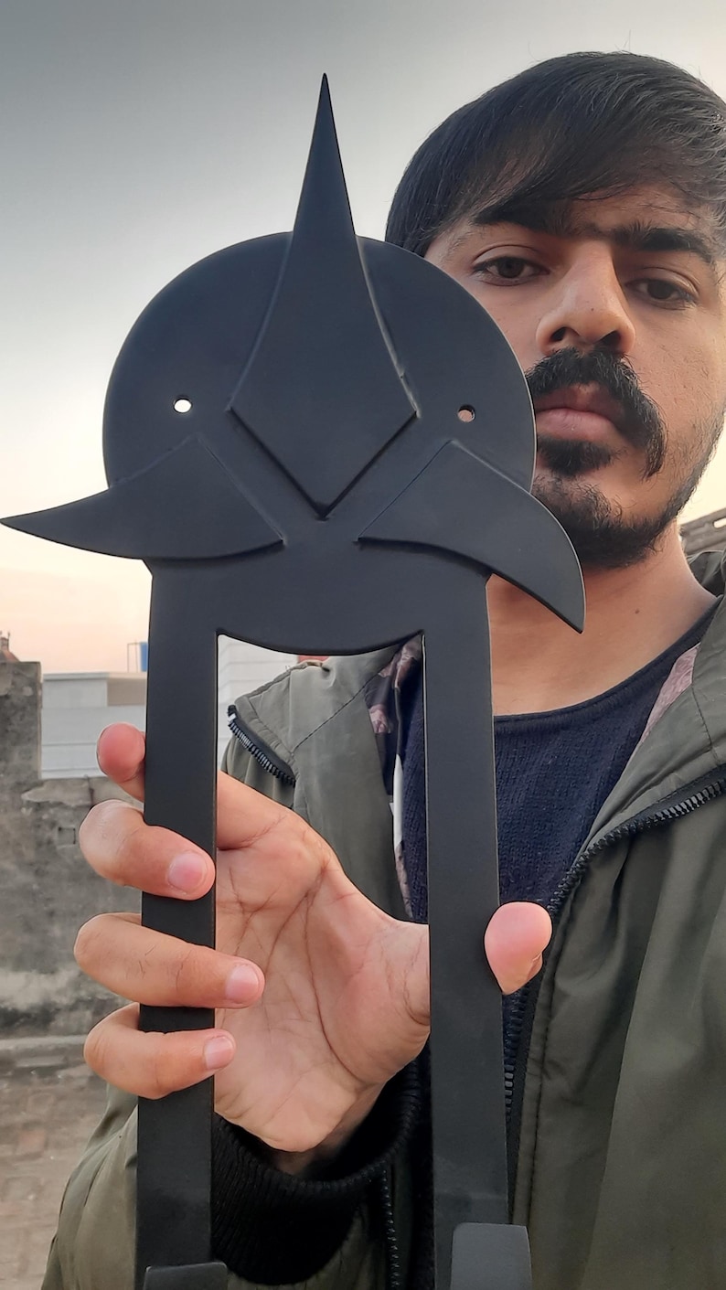 May include: A black metal decorative object with a geometric design. The object has a circular top with a star-like shape and two long, parallel bars extending downwards. The object is held in front of a person.