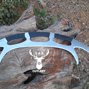 May include: A silver-colored, curved blade with three black-wrapped handles. The blade has a unique design with pointed ends. The logo "SAR CUTLERY" is visible in white. The item is displayed on a weathered wooden surface.