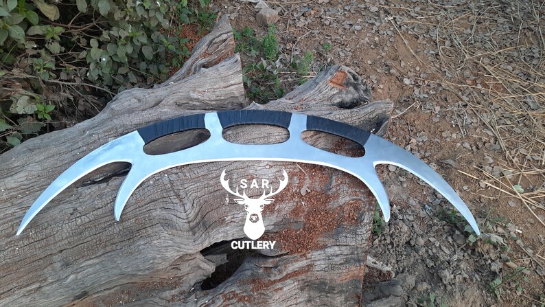 May include: A silver-colored, curved blade with three black-wrapped handles. The blade has a unique design with pointed ends. The logo "SAR CUTLERY" is visible in white. The item is displayed on a weathered wooden surface.