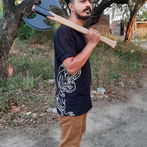 Puede incluir: Una camiseta negra con un estampado gr&aacute;fico blanco, pantalones color canela y zapatillas deportivas negras. Un hacha de batalla de doble cabeza con una cabeza negra mate y un mango de madera clara descansa sobre el hombro. El hacha tiene hojas curvas.