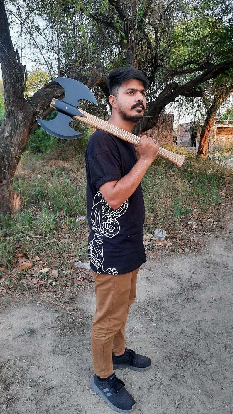 Puede incluir: Una camiseta negra con un estampado gr&aacute;fico blanco, pantalones color canela y zapatillas deportivas negras. Un hacha de batalla de doble cabeza con una cabeza negra mate y un mango de madera clara descansa sobre el hombro. El hacha tiene hojas curvas.