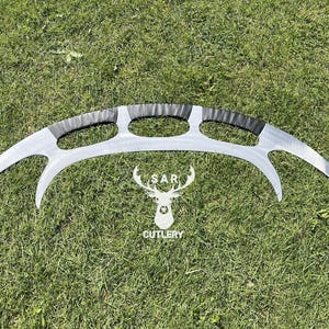 May include: A silver-colored, curved blade with a black handle and three oval cutouts. The blade has a sharp, crescent shape. The logo of a deer head with the text "SAR CUTLERY" is below the blade.