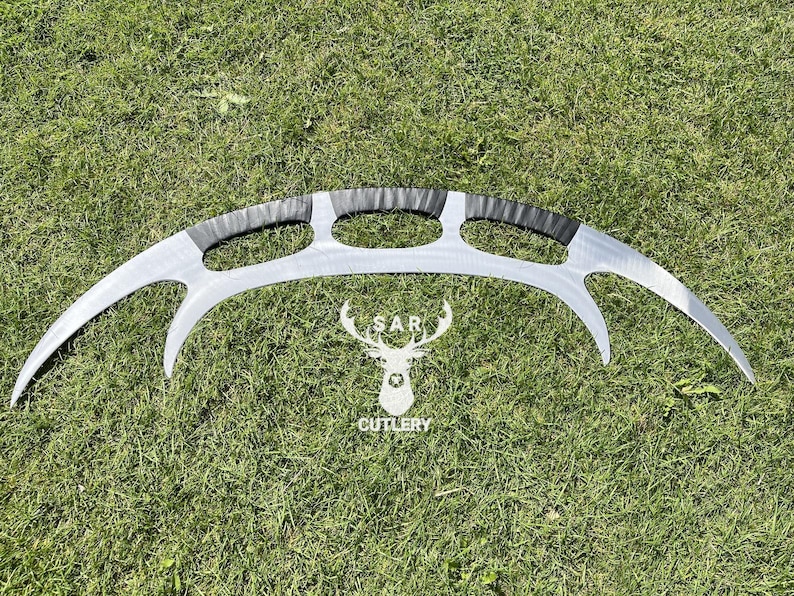 May include: A silver-colored, curved blade with a black handle and three oval cutouts. The blade has a sharp, crescent shape. The logo of a deer head with the text "SAR CUTLERY" is below the blade.