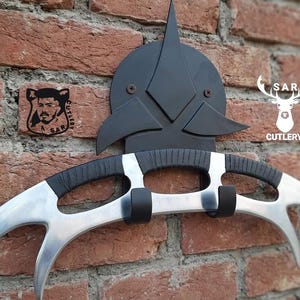 May include: A silver and black Klingon Bat'leth weapon mounted on a brick wall. The curved blade has three points. The handle is black with silver accents. A black emblem is mounted above the weapon. The text "SAR CUTLERY" is visible.