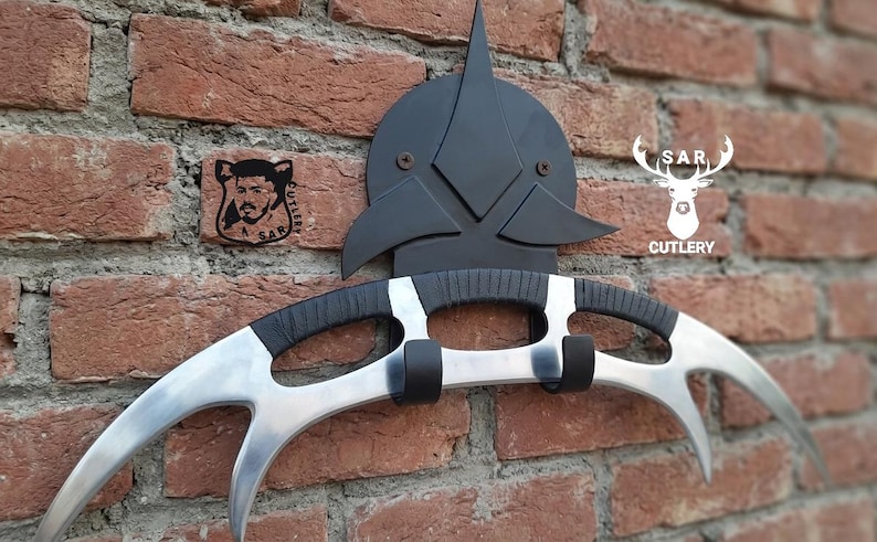 May include: A silver and black Klingon Bat'leth weapon mounted on a brick wall. The curved blade has three points. The handle is black with silver accents. A black emblem is mounted above the weapon. The text "SAR CUTLERY" is visible.