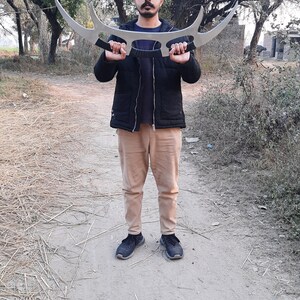 May include: A large, silver-colored, curved blade weapon with two handles. The weapon is held by a person standing outdoors. The person is wearing a black jacket, a blue shirt, tan pants, and black shoes. The background includes trees and a dirt path.