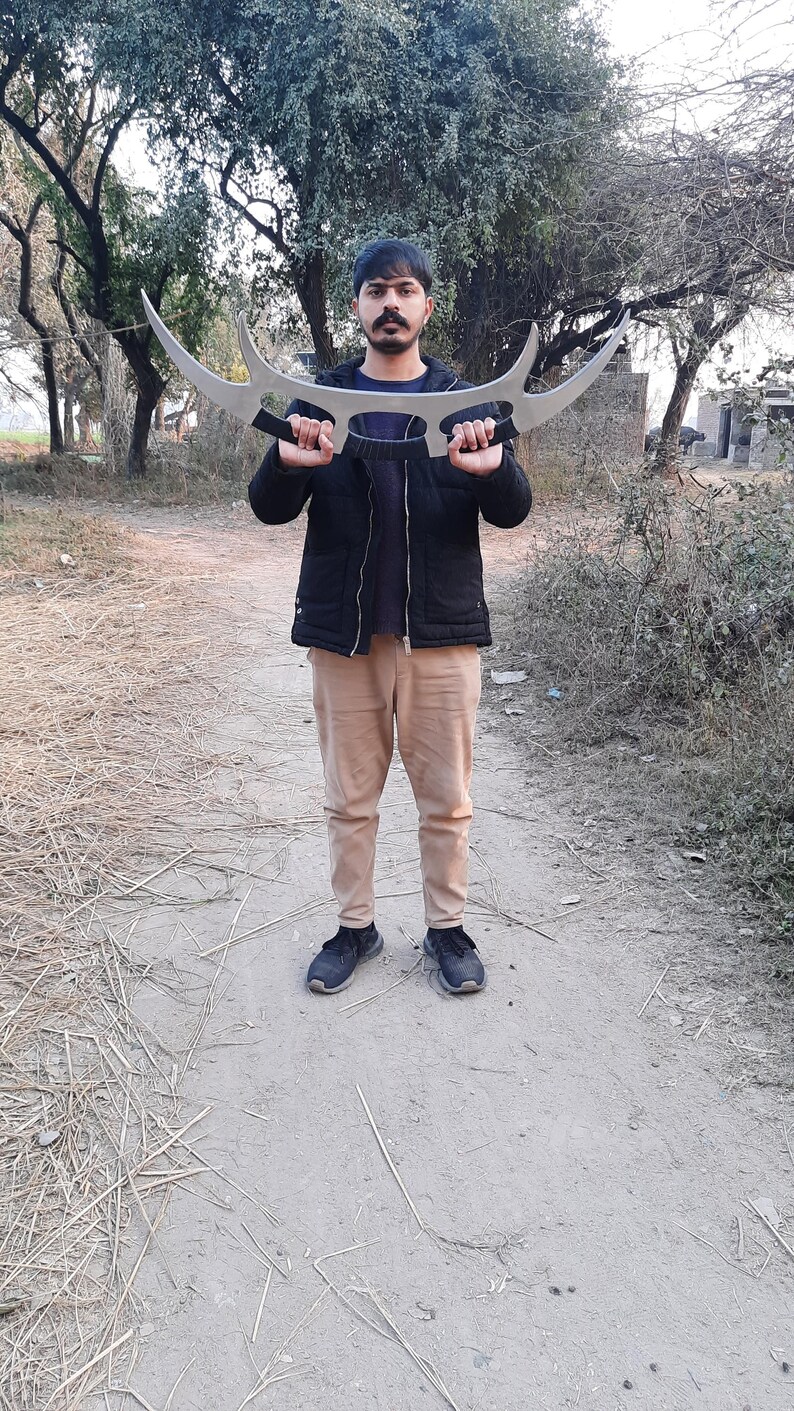 May include: A large, silver-colored, curved blade weapon with two handles. The weapon is held by a person standing outdoors. The person is wearing a black jacket, a blue shirt, tan pants, and black shoes. The background includes trees and a dirt path.