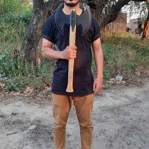 Puede incluir: Un hacha de batalla de doble filo con una cabeza de metal negro mate con dos hojas curvas y un mango de madera largo, recto y de color claro. El hacha se sostiene verticalmente, mostrando el dise&ntilde;o sim&eacute;trico de la hoja y la veta de la madera.