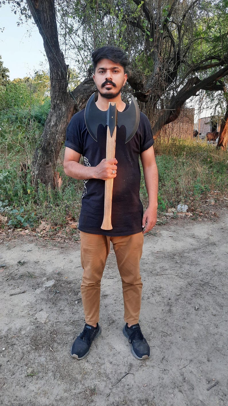Puede incluir: Un hacha de batalla de doble filo con una cabeza de metal negro mate con dos hojas curvas y un mango de madera largo, recto y de color claro. El hacha se sostiene verticalmente, mostrando el dise&ntilde;o sim&eacute;trico de la hoja y la veta de la madera.