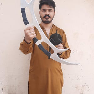 May include: A silver-colored, curved prop weapon with black grips. The prop has a crescent shape with sharp points. A black, circular emblem with a pointed design is also visible.