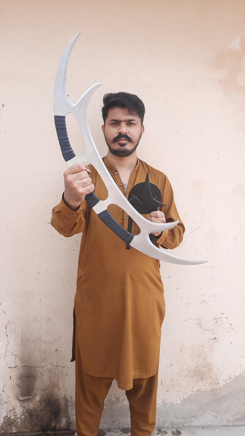 May include: A silver-colored, curved prop weapon with black grips. The prop has a crescent shape with sharp points. A black, circular emblem with a pointed design is also visible.