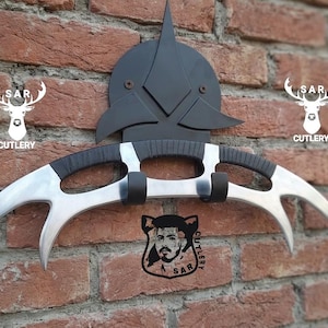 May include: A silver and black curved blade weapon with a black handle, mounted on a black decorative piece. The weapon is displayed against a brick wall, with the text "SAR CUTLERY" and a deer logo.