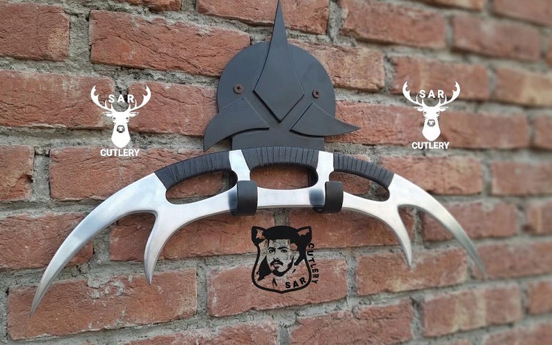 May include: A silver and black curved blade weapon with a black handle, mounted on a black decorative piece. The weapon is displayed against a brick wall, with the text "SAR CUTLERY" and a deer logo.