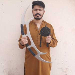 May include: A person holding two props: a large, silver, curved blade with black grips and a black, circular emblem with a pointed design. The props appear to be from a science fiction series.