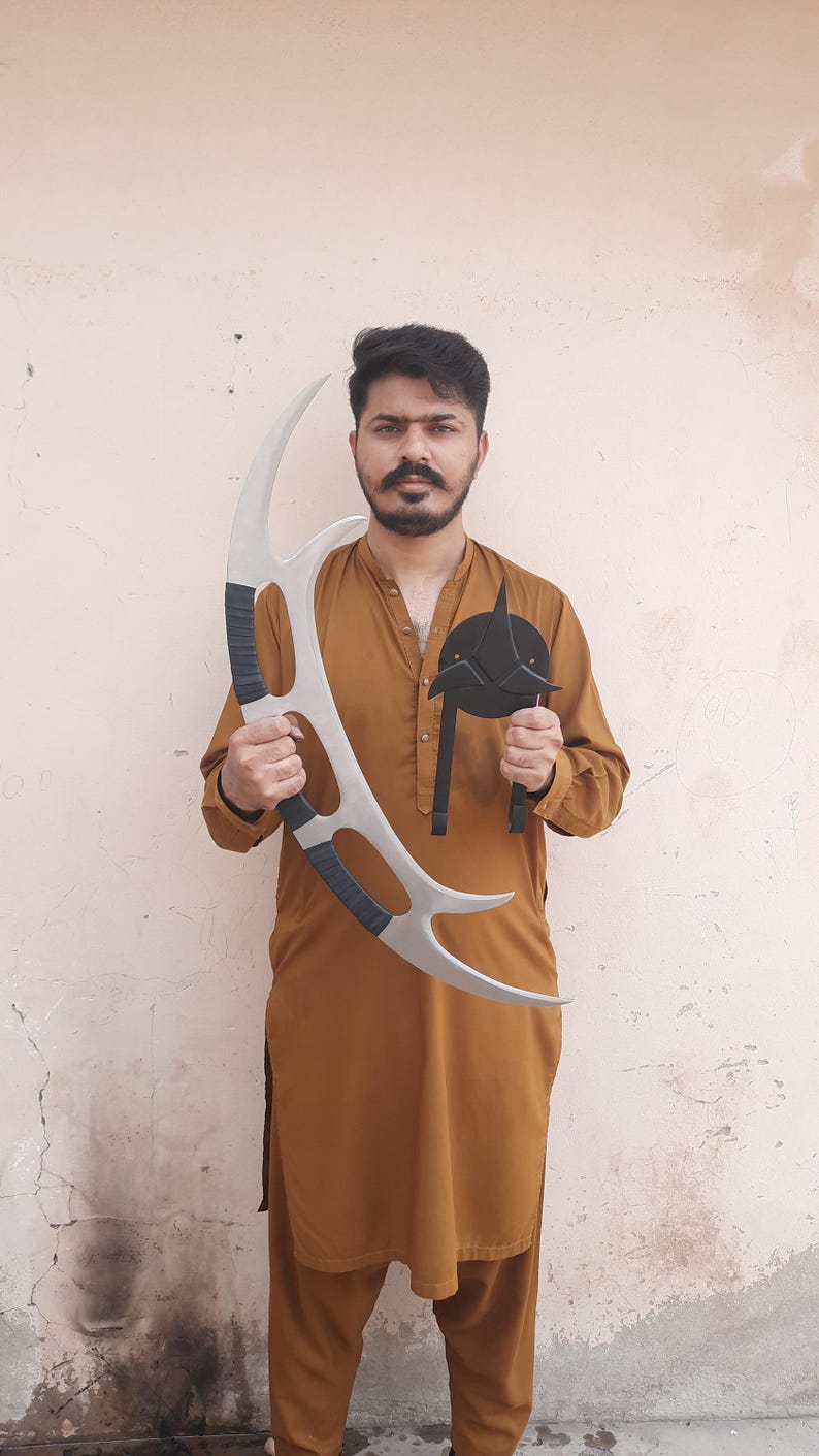 May include: A person holding two props: a large, silver, curved blade with black grips and a black, circular emblem with a pointed design. The props appear to be from a science fiction series.