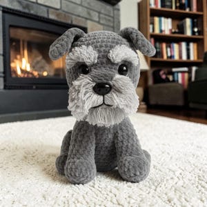 May include: A handmade crochet Schnauzer dog figurine in shades of gray and white. The plush toy has a black nose, large black eyes, and a fluffy beard and eyebrows. The dog is sitting upright on a white rug.