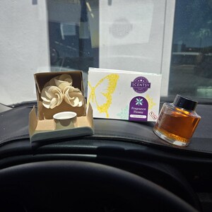 May include: A Scentsy Fragrance Flower set featuring a white paper flower in a small cardboard box, a rectangular product box with a yellow butterfly graphic and purple logo, and a glass bottle filled with amber-colored fragrance oil.