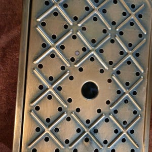 May include: A rectangular brass-coloured metal tray featuring a grid design of intersecting lines and evenly spaced circular perforations. The tray has a raised border and a central circular aperture. Likely for decorative or functional purposes.