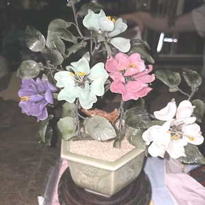 Puede incluir: Una escultura decorativa de árbol bonsái con flores de piedra tallada en tonos morado, verde pálido, rosa y blanco con centros amarillos. El árbol tiene hojas verde oscuro y ramas de alambre, en una maceta hexagonal de cerámica verde claro sobre una base de madera oscura.