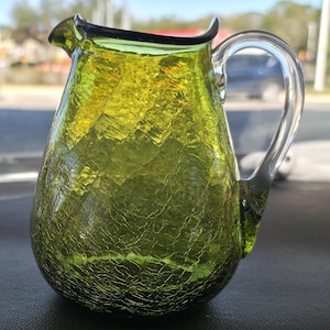 Puede incluir: Una jarra de vidrio verde con acabado craquelado. La jarra tiene un asa curva y un pico vertedor. El vidrio es de un color verde vibrante, y el efecto craquelado añade textura. La jarra se utiliza probablemente para servir bebidas.