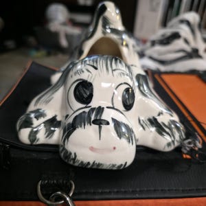 May include: A ceramic dog figurine with a white base and black markings. The dog has large, expressive eyes and a small red mouth. The figurine is lying down, with its head and paws visible. The dog is resting on a black surface.