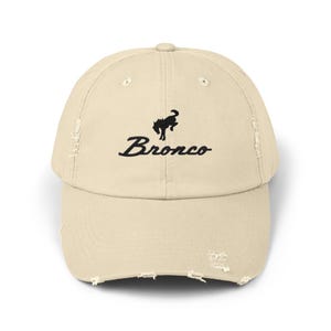 Könnte beinhalten: Beige Baseball Cap mit Used-Look, mit dem Wort "Bronco" in Schreibschrift und einem schwarzen Pferdelogo. Die Kappe hat ein abgenutztes Aussehen mit ausgefransten Kanten.