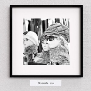 Puede incluir: Fotografía en blanco y negro de tres cabezas de maniquíes con sombreros, gafas de sol y bufandas de piel sintética. La imagen está enmarcada en un marco cuadrado negro y montada en una pared blanca. El texto "The Gossips - 2009" está en la parte inferior.