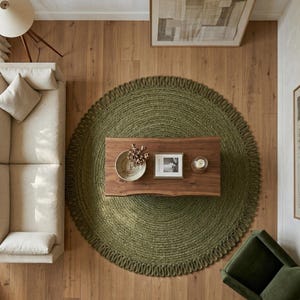 May include: A large, round, olive green braided jute rug with a thick, looped edge texture. The rug is positioned centrally on a light wood floor, serving as a base for a rectangular wooden coffee table with a natural live edge finish.