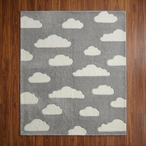 May include: A rectangular gray rug with white cloud shapes scattered across its surface. The rug has a soft, plush texture, and the clouds are slightly raised, creating a three-dimensional effect. The rug is designed for a nursery or children's room.