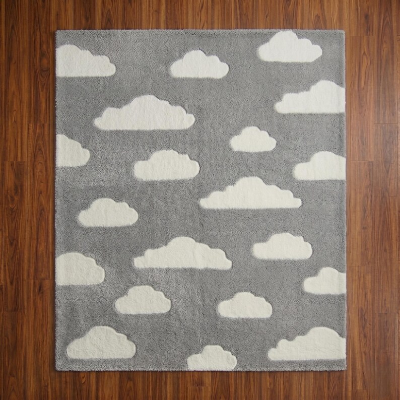 May include: A rectangular gray rug with white cloud shapes scattered across its surface. The rug has a soft, plush texture, and the clouds are slightly raised, creating a three-dimensional effect. The rug is designed for a nursery or children's room.