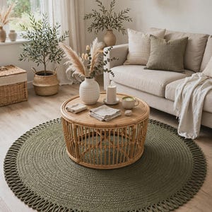 May include: A round, olive green braided rug featuring a decorative looped edge border. The rug is constructed from a thick, textured cord material and is positioned on a light-coloured wood floor beneath a circular rattan coffee table.