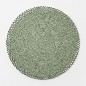 May include: A round sage green placemat featuring a tightly coiled braided centre and a decorative looped edge border. The placemat has a uniform circular shape and a textured surface created by the woven material, providing a consistent colour.
