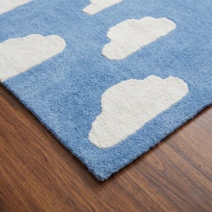 May include: A blue rug with white cloud shapes. The rug has a soft, textured surface. The clouds are fluffy and three-dimensional, creating a whimsical design. The rug is placed on a wooden floor.