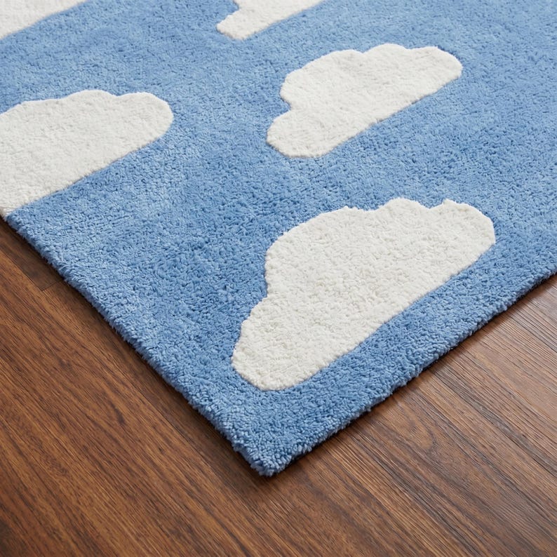 May include: A blue rug with white cloud shapes. The rug has a soft, textured surface. The clouds are fluffy and three-dimensional, creating a whimsical design. The rug is placed on a wooden floor.