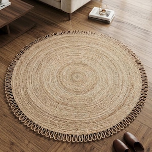 May include: A round, tan-colored woven jute rug featuring a concentric circular pattern and a decorative looped fringe border. The rug is constructed from natural fibers, showcasing a textured, braided surface design throughout the entire piece.