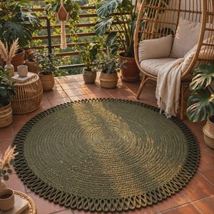 May include: A circular, olive green area rug featuring a thick, braided texture throughout the centre and a decorative, looped border design. The rug is crafted from a heavy, cord-like material and lies flat on a tiled floor surface.