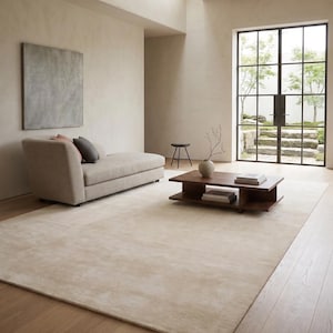 May include: A neutral-toned living room featuring a large, light-colored rug, a low wooden coffee table, and a beige chaise lounge. A large window offers a view of an outdoor garden. The room is decorated in a minimalist style.