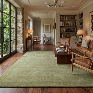 May include: A large, rectangular area rug in a solid sage green color with a subtle border detail. The rug is positioned on a dark wood floor in a living room setting, featuring a low-pile texture that covers a significant portion of the floor space.