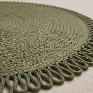 May include: A circular olive green braided mat featuring a textured woven surface and a decorative looped fringe border. The mat is constructed from thick, tightly coiled fibres that create a consistent pattern across the entire round shape.