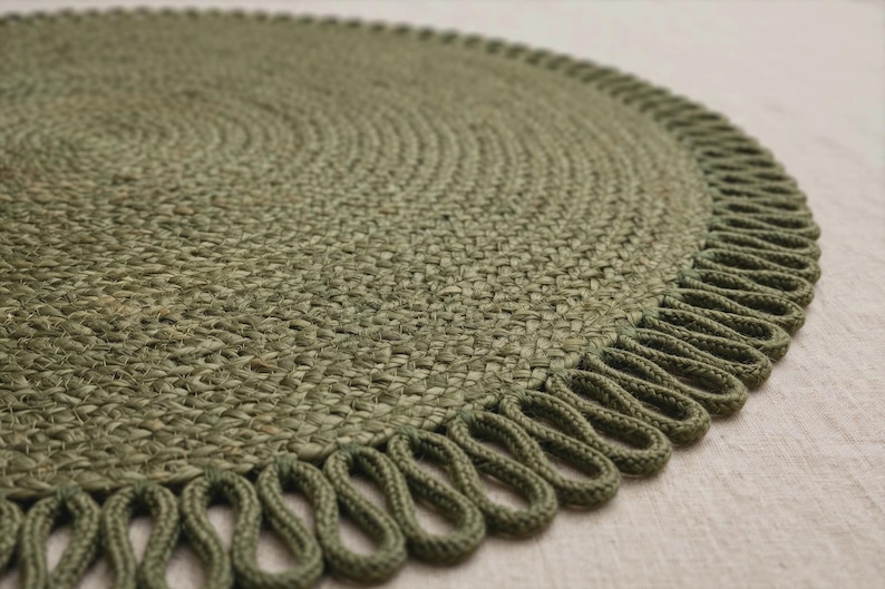 May include: A circular olive green braided mat featuring a textured woven surface and a decorative looped fringe border. The mat is constructed from thick, tightly coiled fibres that create a consistent pattern across the entire round shape.