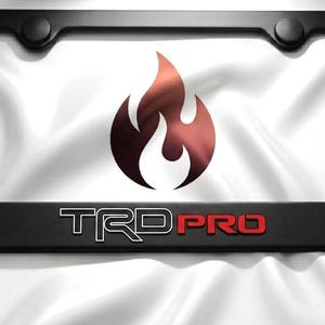 May include: Black license plate frame with the text "TRD PRO" in white and red. The frame features a flame graphic in the center. The background is a white, silky fabric.