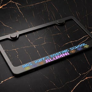 May include: A black rectangular license plate frame featuring the text "MRS. PUFF'S BOATING SCHOOL ALUMNI" in blue and pink lettering. The frame is decorated with small, colorful flower icons in yellow, orange, and purple along the bottom edge.