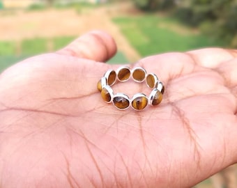 May include: A silver ring with oval tiger's eye stones set around the band. The ring is held in a hand, with a blurred green background.